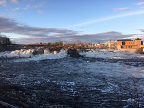 A scenic view of a waterfall with rushing water, surrounded by buildings and a blue sky with clouds.