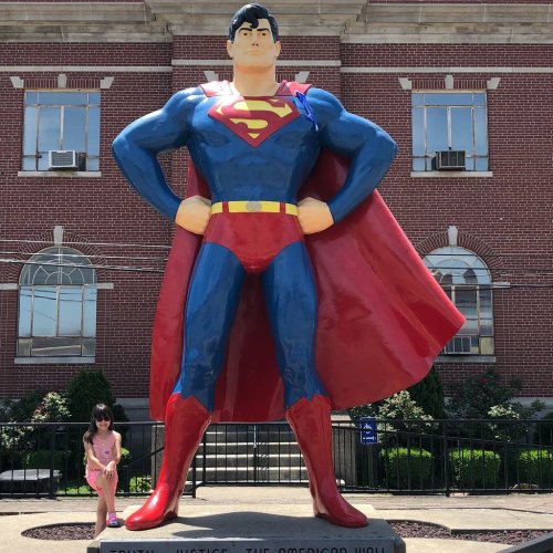 A young girl stands beside a large Superman statue in front of a brick building.