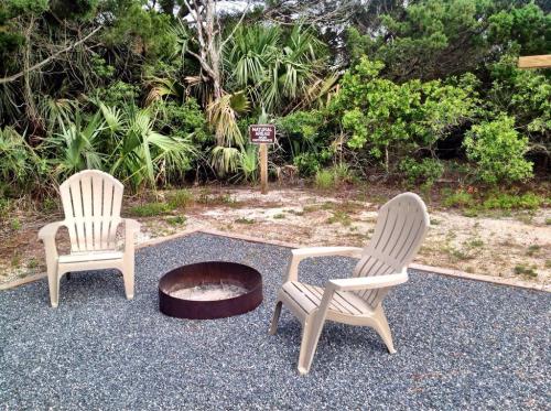 Two beige chairs sit beside a circular fire pit on gravel, surrounded by lush greenery and a sign in the background.