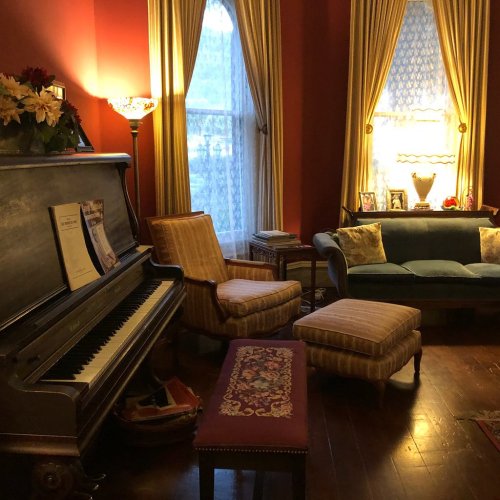 Cozy living room with a piano, vintage furniture, and warm lighting, featuring red walls and large windows.