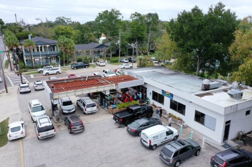 Aerial view of a busy restaurant with outdoor seating, surrounded by parked cars and trees in a residential area.