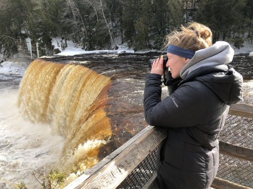 A person in a black jacket takes photos of a waterfall from a wooden railing, surrounded by trees.