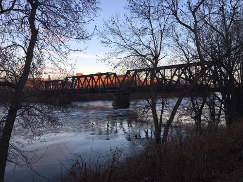 A rusted train bridge spans a river, framed by bare trees and a colorful sunset in the background.