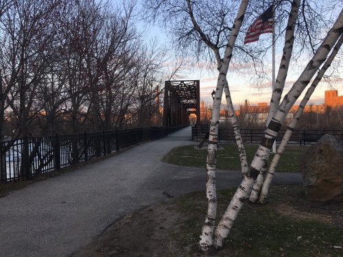 A pathway leads to a bridge, framed by trees and an American flag, with a sunset in the background.