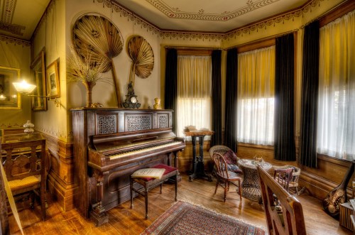 A vintage room featuring a piano, elegant furniture, and decorative fans on the wall, with soft natural light filtering through curtains.