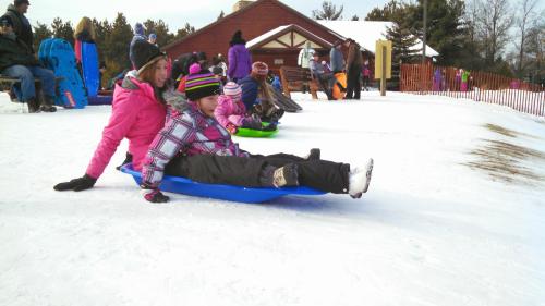A woman and a child sit on a sled, ready to go down a snowy hill, with others in the background enjoying winter activities.