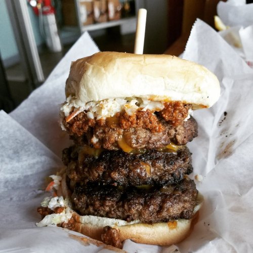 A towering burger with three beef patties, cheese, coleslaw, and a top bun, served on parchment paper.