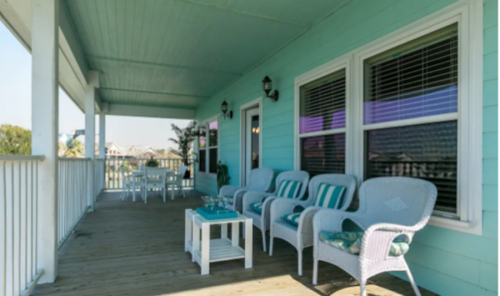 A light blue porch with white wicker chairs, a small table, and a dining set, surrounded by a sunny outdoor view.