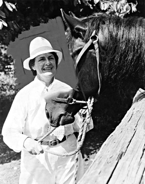 A smiling person in a white outfit holds a horse's reins, standing beside a horse in a natural setting.