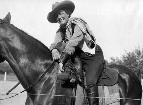 A smiling cowboy in vintage attire sits on a horse, adjusting his boot with a scenic background.