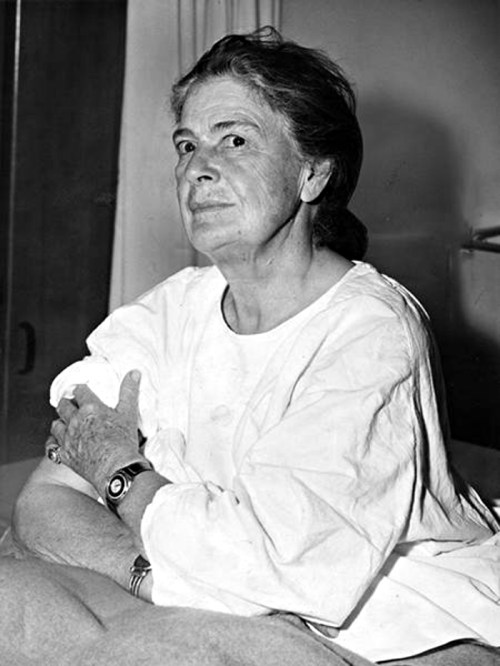 A woman in a white shirt sits on a bed, looking thoughtfully to the side with a serious expression.