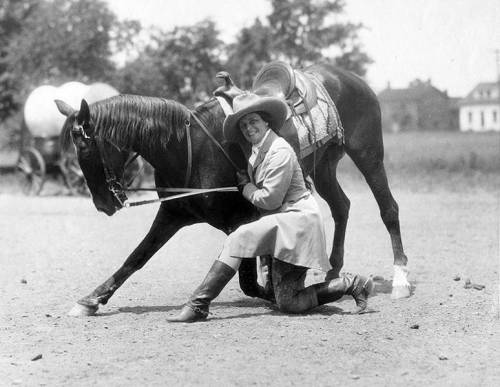 A woman in a hat kneels beside a horse, smiling as she interacts with it in a vintage outdoor setting.