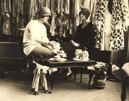 Two women in vintage clothing sit at a table with tea and pastries, surrounded by dresses and a decorative stuffed animal.