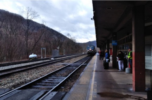 A train station platform with passengers waiting as a train approaches, surrounded by mountains and cloudy skies.