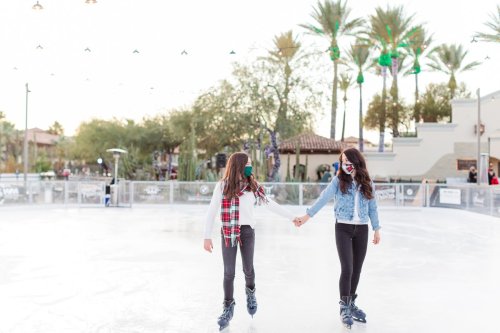Two girls ice skating hand in hand on an outdoor rink, surrounded by palm trees and festive decorations.