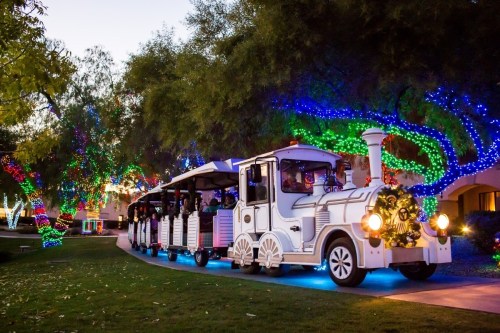 A festive white train decorated with lights, surrounded by colorful illuminated trees in a park setting.