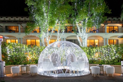 A cozy, illuminated igloo structure surrounded by twinkling lights and trees at night, with a hotel in the background.