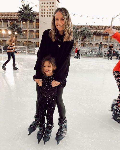 A woman and a young girl smile while ice skating together on a rink, surrounded by festive decorations.