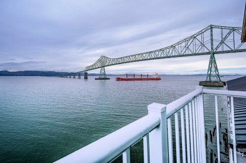 A large bridge spans a calm waterway, with a cargo ship passing underneath and a railing in the foreground.