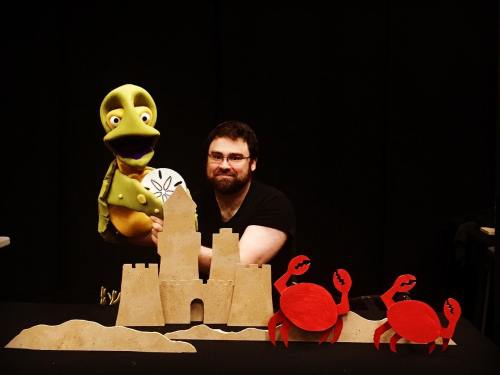 A puppeteer holds a turtle puppet in front of a sandcastle and red crab cutouts on a black background.