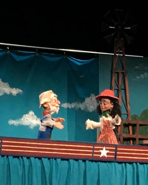 Two puppets on stage: an elderly man with a beard and a woman in a red hat, set against a colorful backdrop.