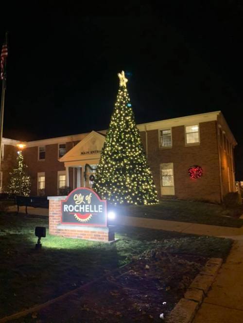 A brightly lit Christmas tree stands in front of the City of Rochelle building, decorated for the holiday season at night.