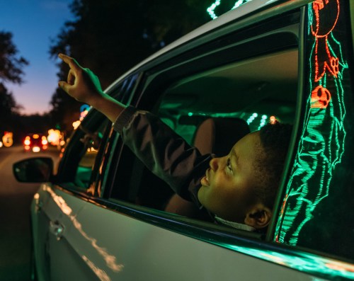 A child leans out of a car window, pointing excitedly at colorful lights in the evening sky.