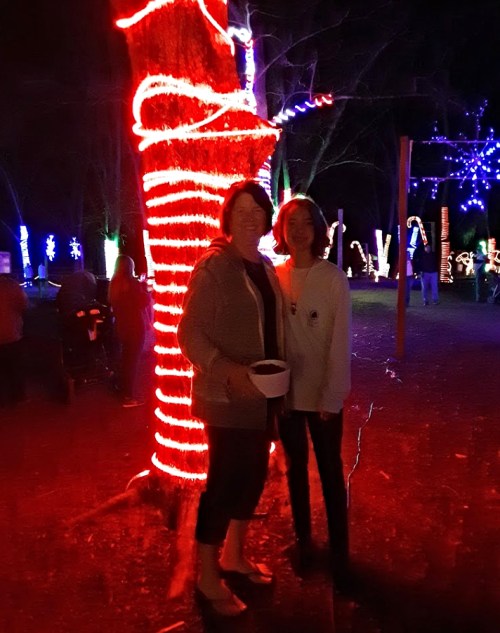 Two people stand together near a brightly lit tree decorated with red lights in a festive outdoor setting.