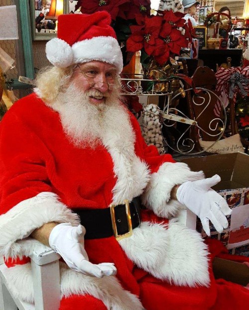 Santa Claus in a red suit and hat, sitting in a festive setting with poinsettias and holiday decorations.
