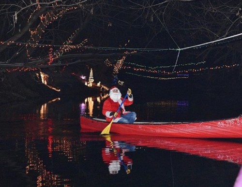 A person dressed as Santa paddles a red canoe on a dark river, surrounded by colorful holiday lights.
