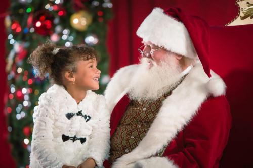 A joyful girl in a white coat smiles at Santa Claus, who is dressed in his traditional red suit, in a festive setting.