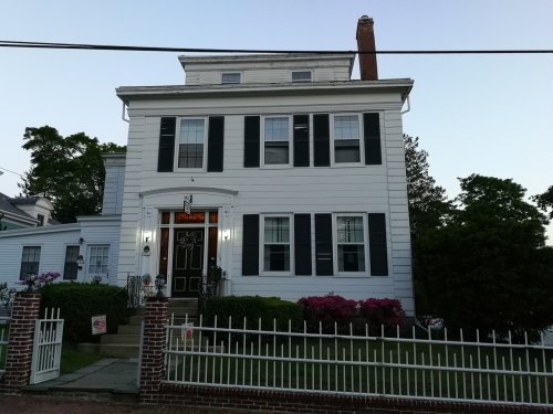 A two-story white house with black shutters, a front porch, and a fenced yard, surrounded by greenery and flowers.