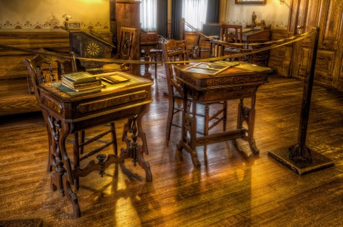 Historic room featuring ornate wooden desks, books, and vintage furniture, illuminated by soft natural light.