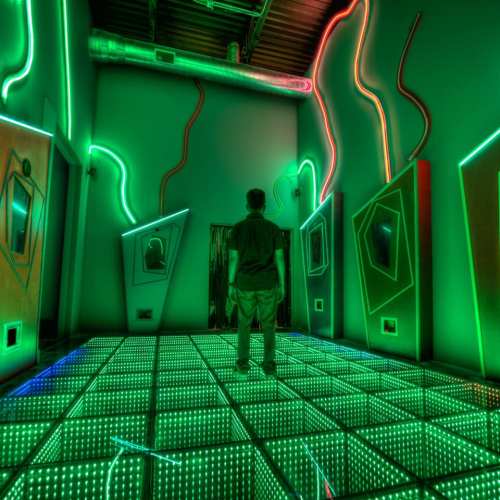 A person stands in a neon-lit room with mirrored floors and glowing geometric shapes on the walls.