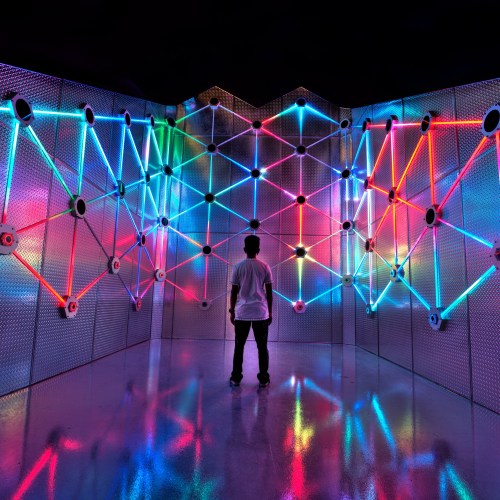A person stands in front of a vibrant, illuminated wall with colorful neon lights and geometric patterns.