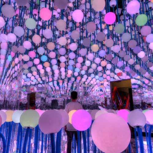 A person stands in a mirrored room filled with colorful, glowing spheres hanging from the ceiling.