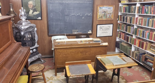 A vintage classroom with a chalkboard, wooden desks, a piano, and shelves filled with books.