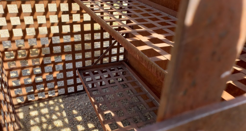 Close-up of a rusty, lattice-patterned metal structure with shelves, casting shadows on a gravel surface.