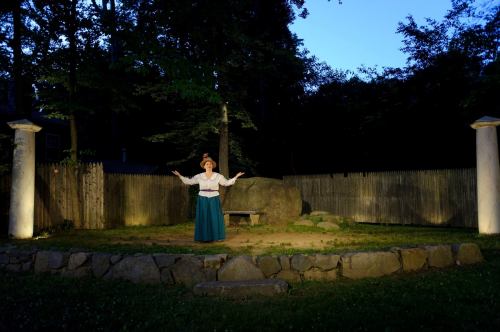 A performer in period costume stands on a stone stage surrounded by trees and illuminated by soft lighting at dusk.