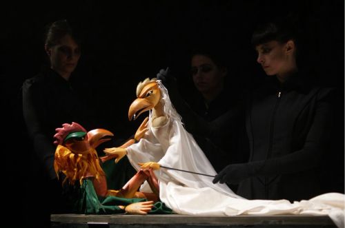 Two puppeteers manipulate colorful bird puppets on a dark stage, with one puppet draped in a white cloth.