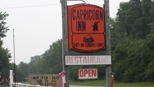 Sign for Capricorn Inn restaurant, featuring "Eat-In Carry-Out" and "Open" banners, surrounded by greenery.