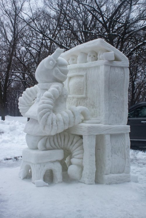 A snow sculpture of a character playing a piano, surrounded by trees and snow.