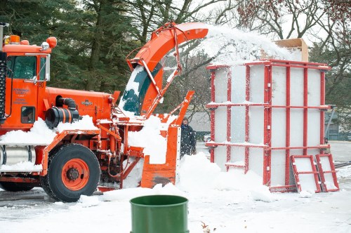 A bright orange snowplow is clearing snow and depositing it into a large red container on a snowy day.