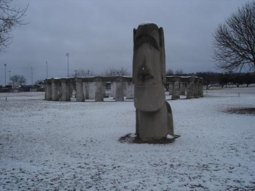 A snow-covered landscape featuring a large stone sculpture resembling a moai, with a stone circle in the background.