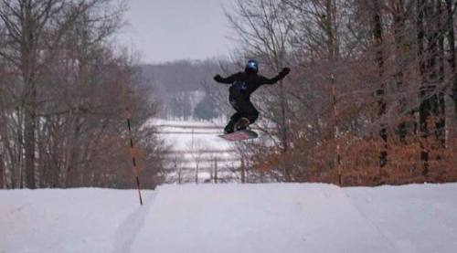A snowboarder jumps off a snowy ramp, surrounded by trees in a winter landscape.