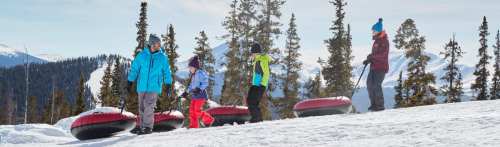 A family walks on a snowy hill with snow tubes, surrounded by trees and mountains under a clear blue sky.