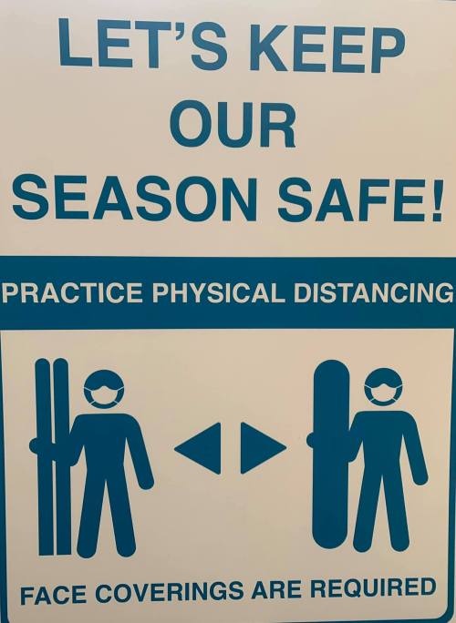 Sign promoting safety measures: "Practice physical distancing" and "Face coverings are required" with icons of skiers.