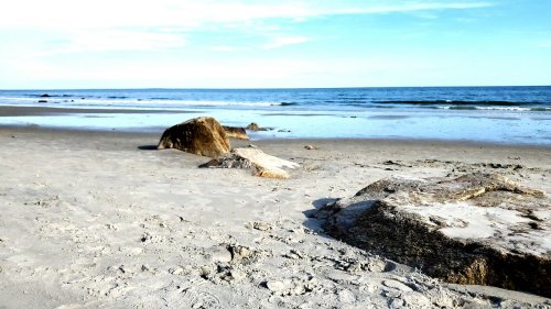 A serene beach scene with smooth sand, scattered rocks, and gentle waves under a clear blue sky.