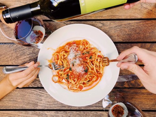 A plate of spaghetti with tomato sauce and cheese, surrounded by wine glasses and hands holding forks.