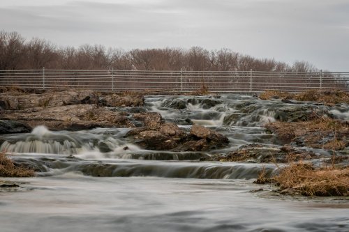 A serene river flows over rocks, surrounded by sparse vegetation and a distant tree line under a cloudy sky.